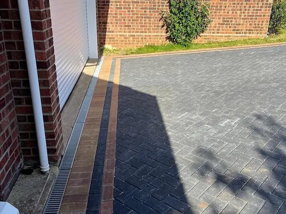 Charcoal herringbone block paving with a buff border and a drainage channel running along a garage wall