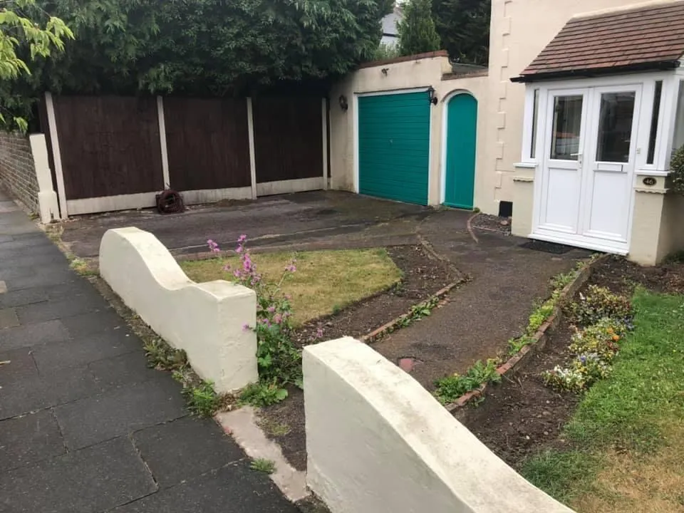 Worn tarmac drive, lawn and brick-edged beds behind curved white walls in front of a green garage door