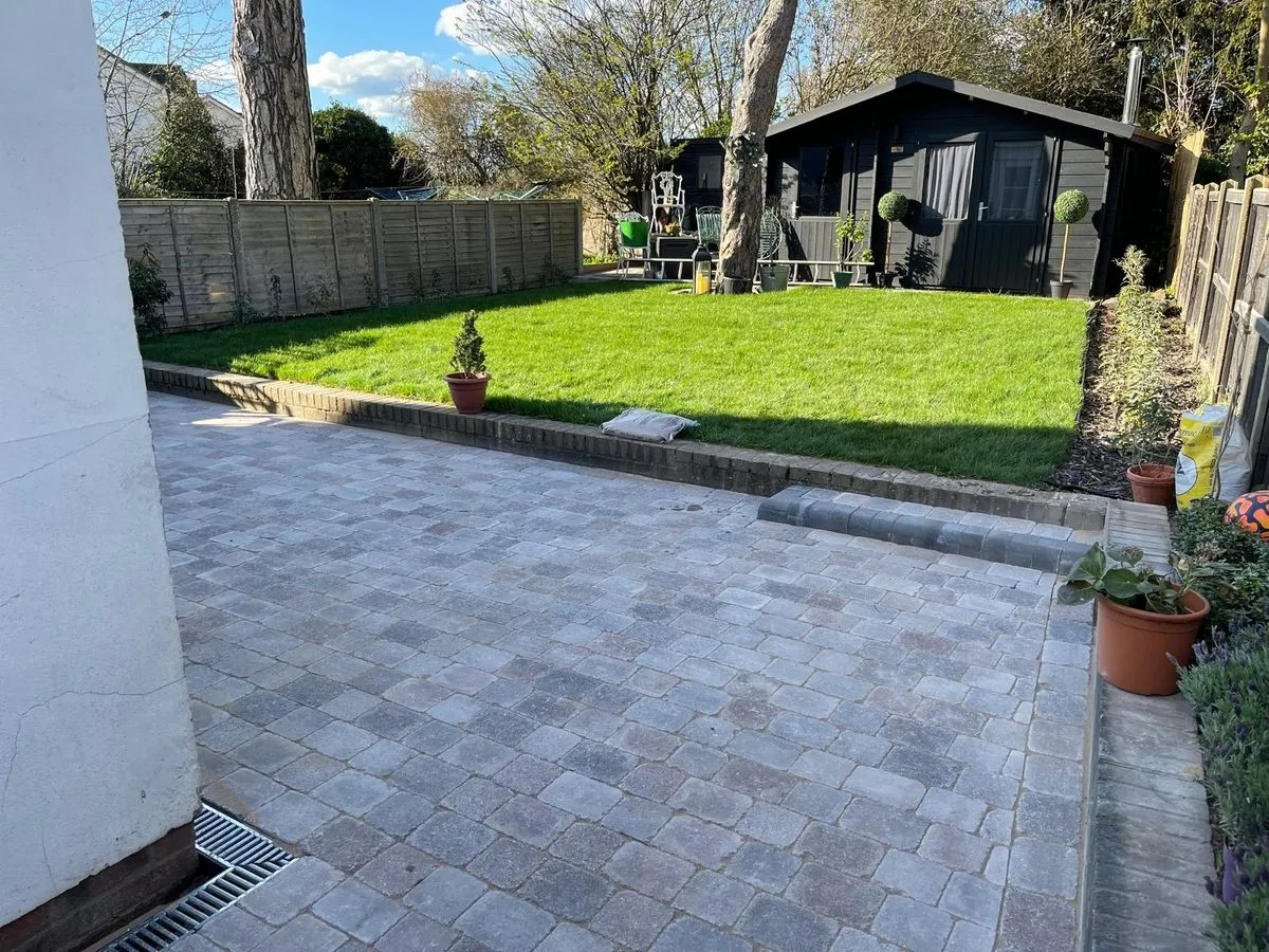 Grey tumbled block paving patio with a lawn and summerhouse behind, laid by First4Driveways